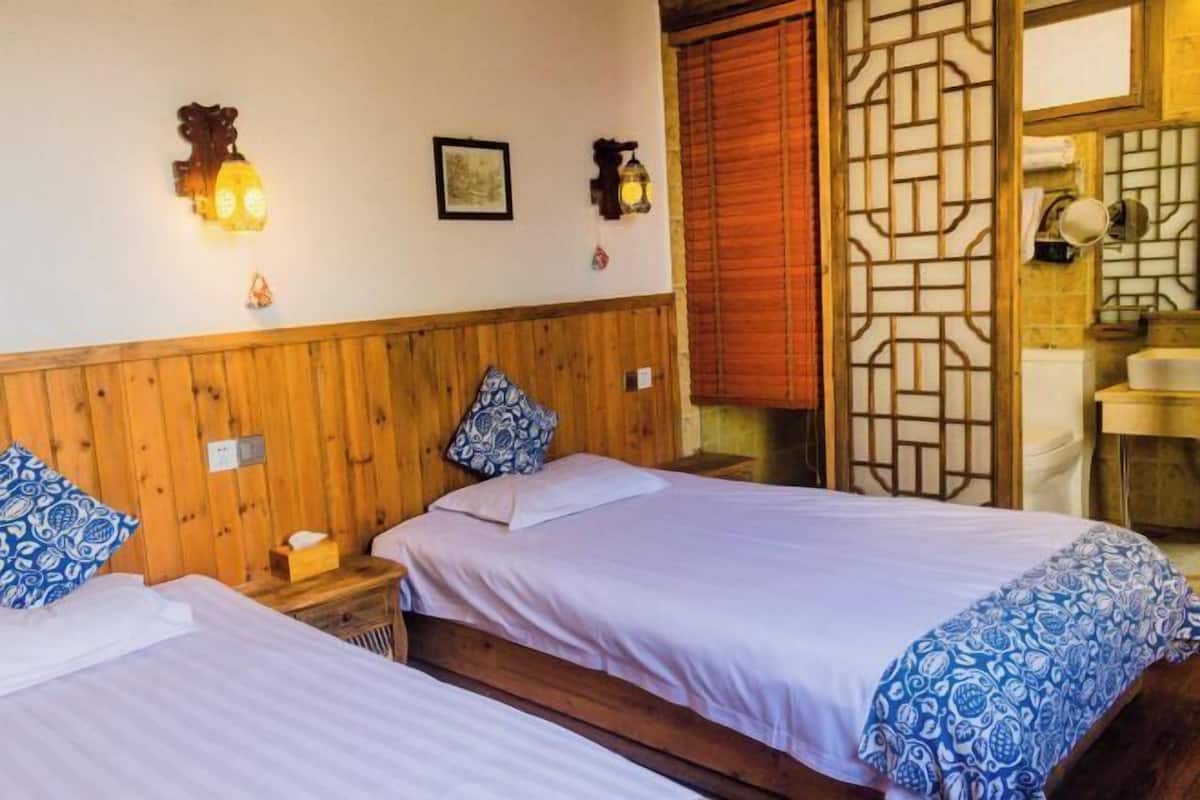 A room with two beds, wooden headboard, wall-mounted lights, a wooden partition, and a small bedside table.