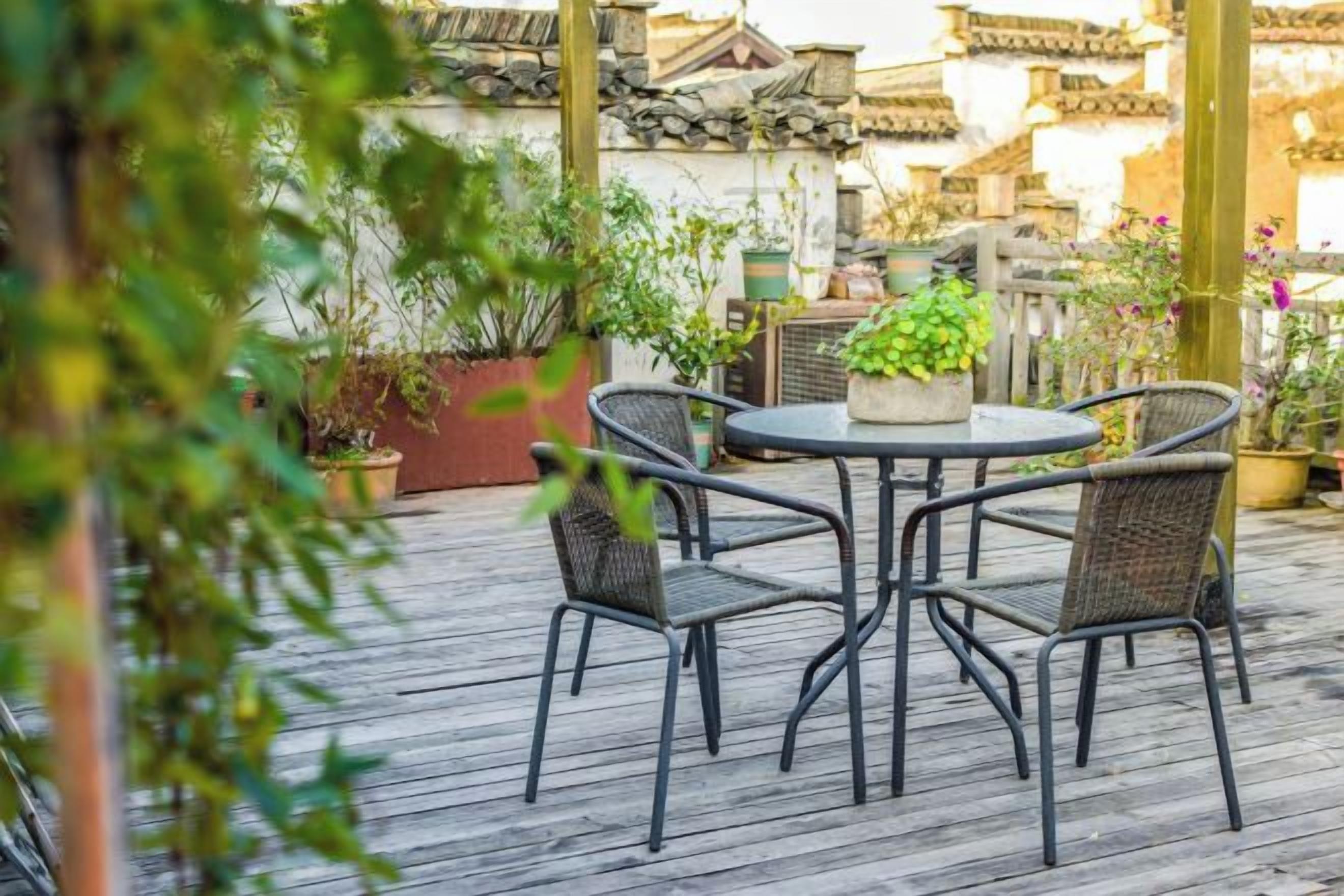 Un patio exterior con una mesa redonda y cuatro sillas, rodeado de plantas en macetas y una terraza de madera.