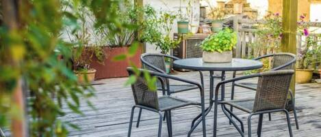 Un patio exterior con una mesa redonda y cuatro sillas, rodeado de plantas en macetas y una terraza de madera.