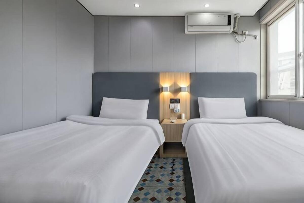 A hotel room with two beds, a nightstand with a lamp, an air conditioning unit, and a window.