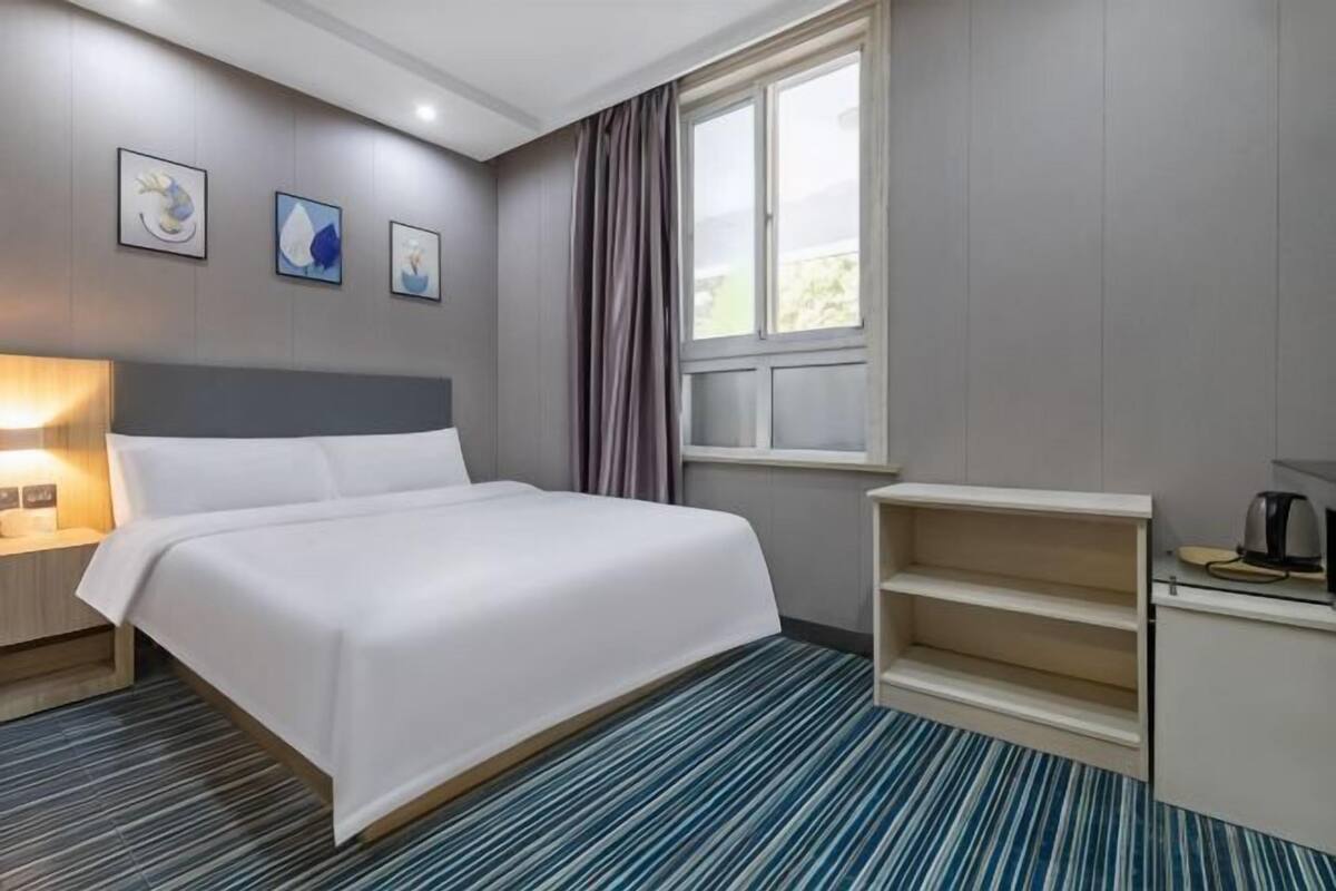 A hotel room with a bed, bedside table, lamp, window with curtains, and a small desk with a kettle.