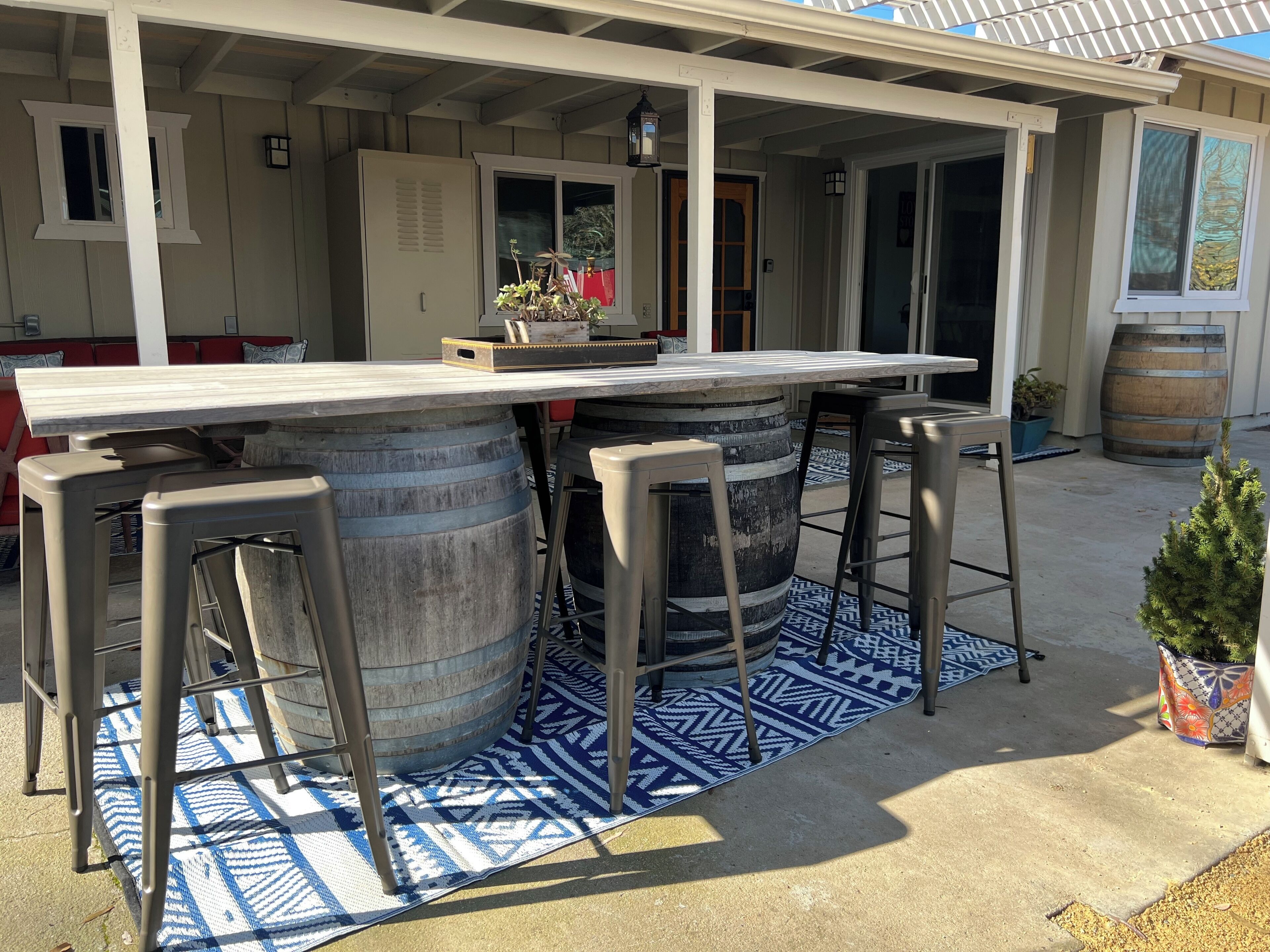 En udendørs patio med et barområde, hvor der er to store tønder som barstole, et langt bord og en farverig måtte.