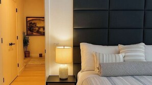 Une chambre à coucher comprenant un lit, une table de chevet avec une lampe, une tête de lit fixée au mur, une porte et un couloir menant à une autre pièce.