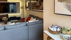 Un salon moderne avec un canapé d’angle gris, une table basse et un téléviseur. Des œuvres d’art encadrées ornent le mur, et une tasse de thé est posée sur la table d’appoint.