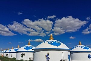 A row of white yurts with blue and gold accents, set against a clear blue sky with scattered clouds.
