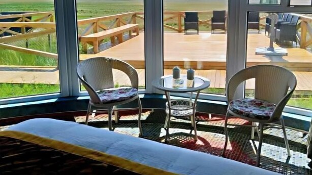 Una habitación con un ventanal grande, una cama, dos sillas de mimbre, una mesita redonda y una terraza de madera afuera.