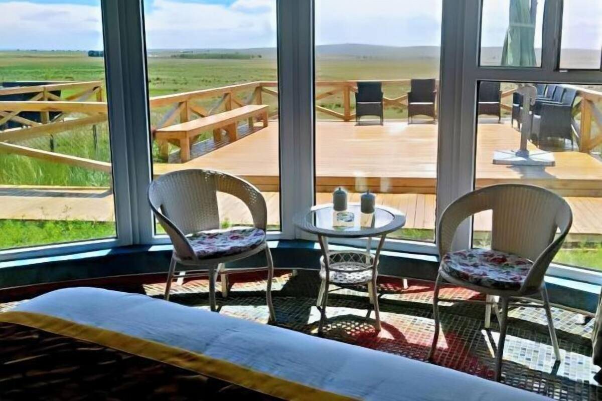 A room with a large window, a bed, two wicker chairs, a small round table, and a wooden deck outside.