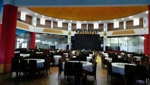 Un salón amplio con varias mesas redondas y sillas, un escenario con una pantalla y un techo alto con un diseño decorativo.