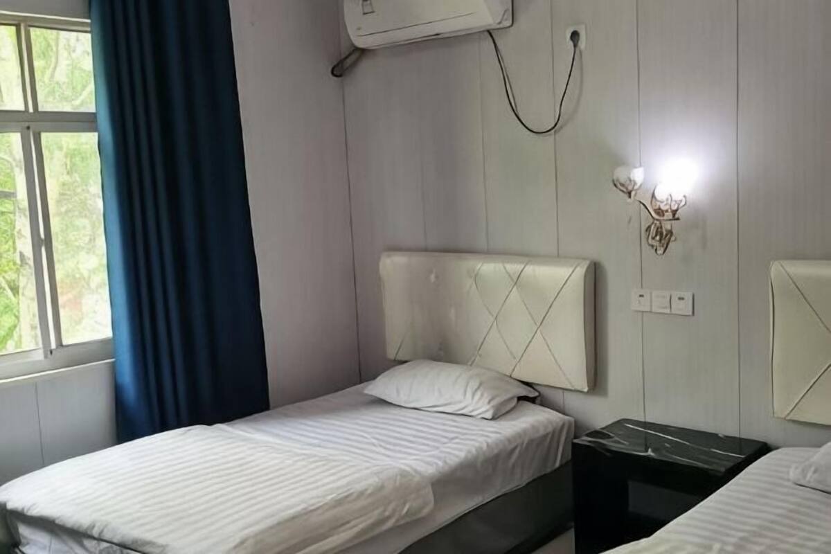 Quarto com duas camas, janela com cortinas, ar condicionado e luminária fixada na parede.