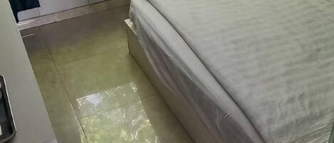 A bed with a white cover, a tiled floor, and a white wall.