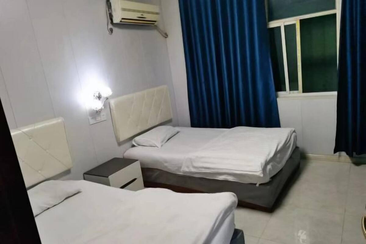 Quarto de hotel com duas camas, mesa de cabeceira, luminária, janela com cortinas e ar condicionado.