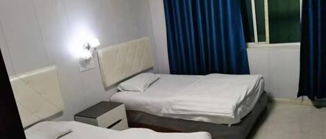 A hotel room with two beds, a bedside table, a lamp, a window with curtains, and an air conditioning unit.