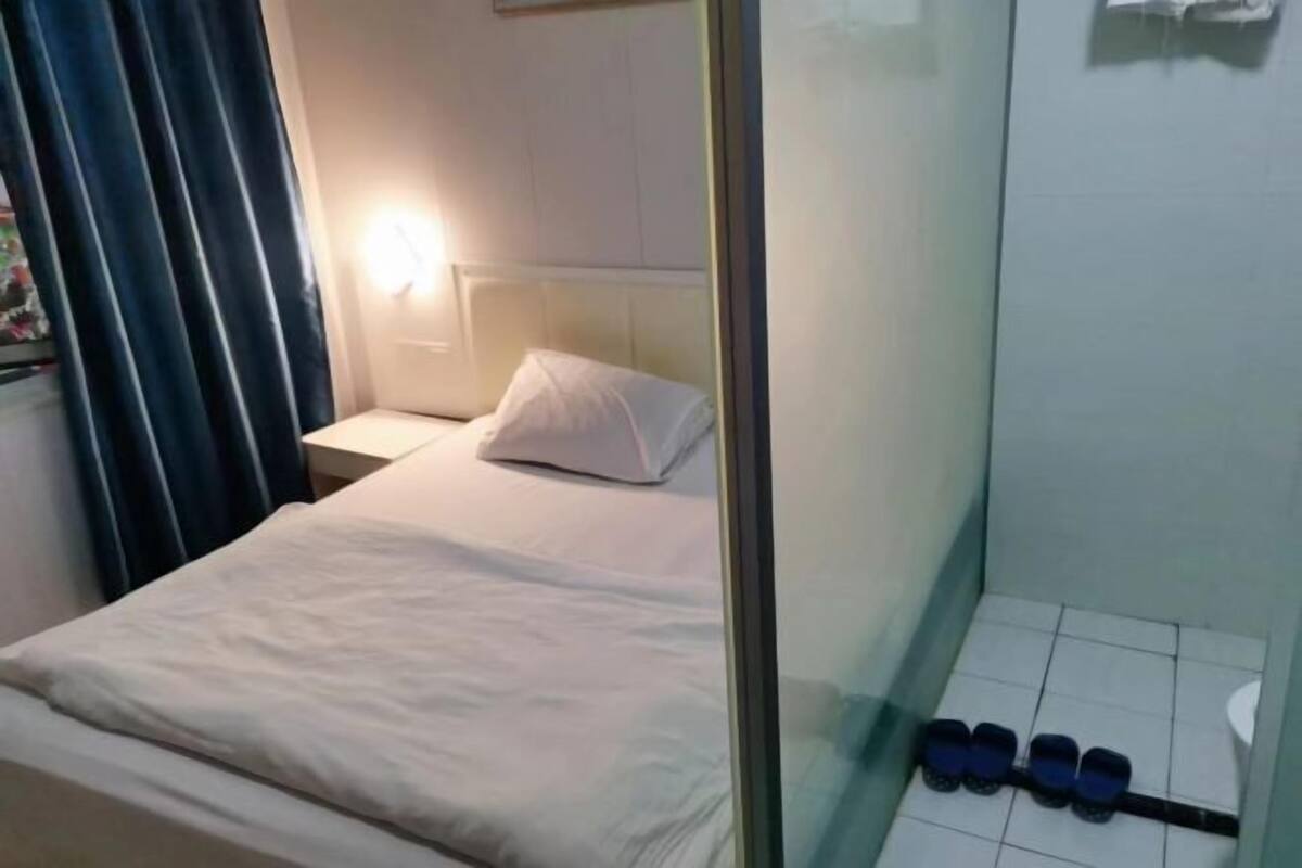 Uma cama de solteiro com roupa de cama branca, uma mesa de cabeceira, uma luminária na parede e uma pequena prateleira com garrafas.