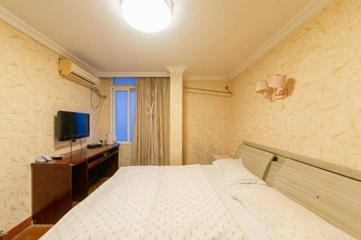 Habitació d'hotel amb un llit, una televisió sobre un suport de fusta, una finestra amb cortines i un aire condicionat fixat a la paret.
