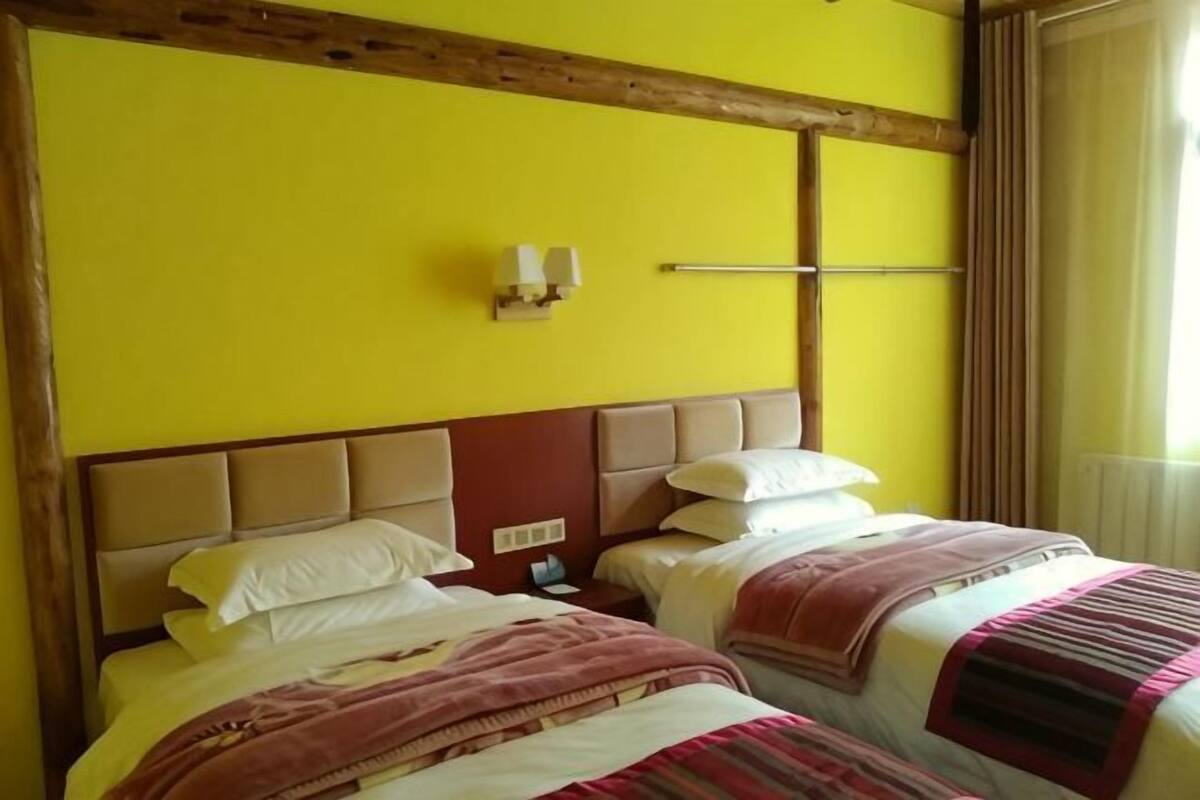 A hotel room with two beds, a wooden ceiling beam, a wall-mounted light fixture, and a window with curtains.