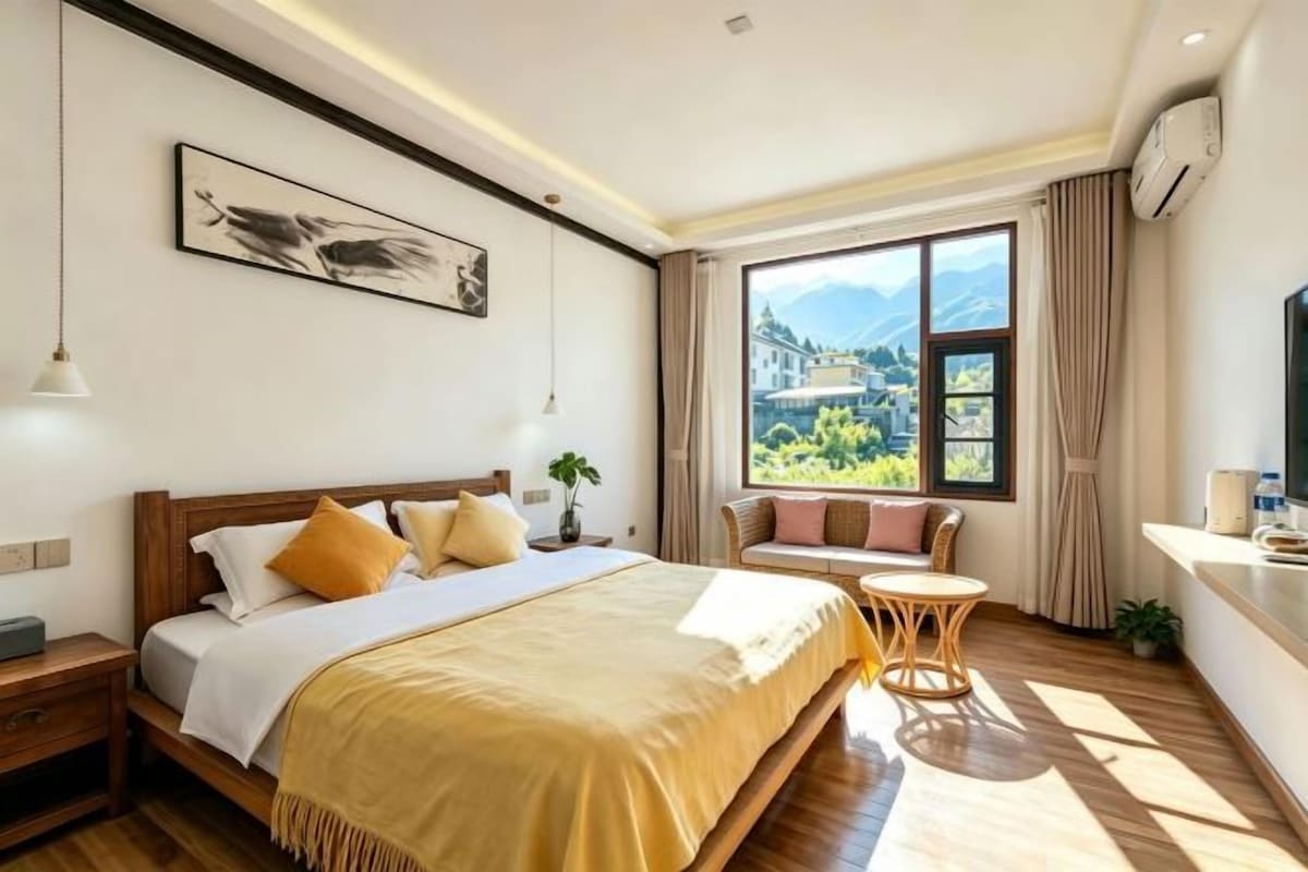 A hotel room with a large bed, a bedside table, a window with a view of mountains, and a small seating area.