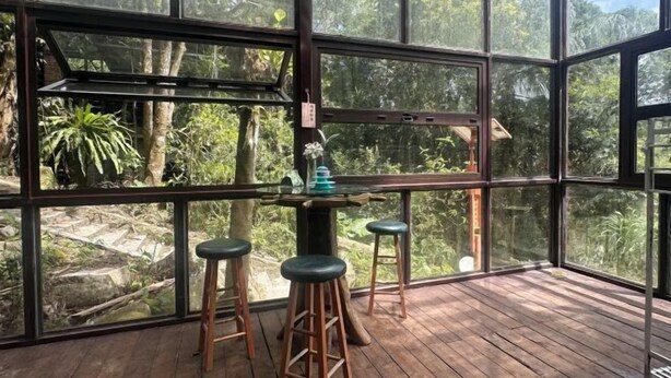 Une pièce dotée de grandes fenêtres donnant sur un paysage verdoyant et une forêt dense. On y trouve une table en bois, des tabourets et un vase rempli de fleurs.