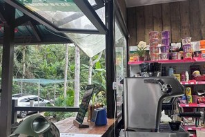 Interior de una cafetería con una máquina de café, estanterías con diversos artículos y vistas a espacios verdes al exterior.