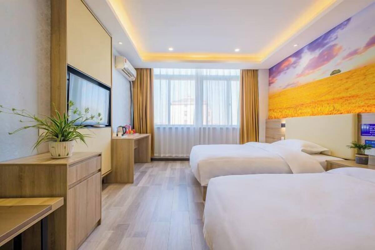 A hotel room with two beds, a TV, a desk, a plant, and a painting on the wall.