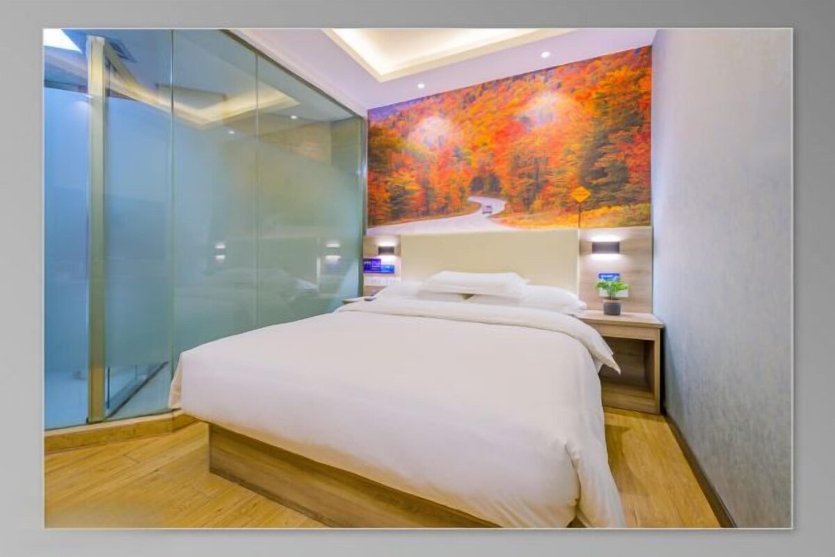 A modern hotel room with a glass partition, a bed with white linens, a wall mural, and a bedside table with a plant.