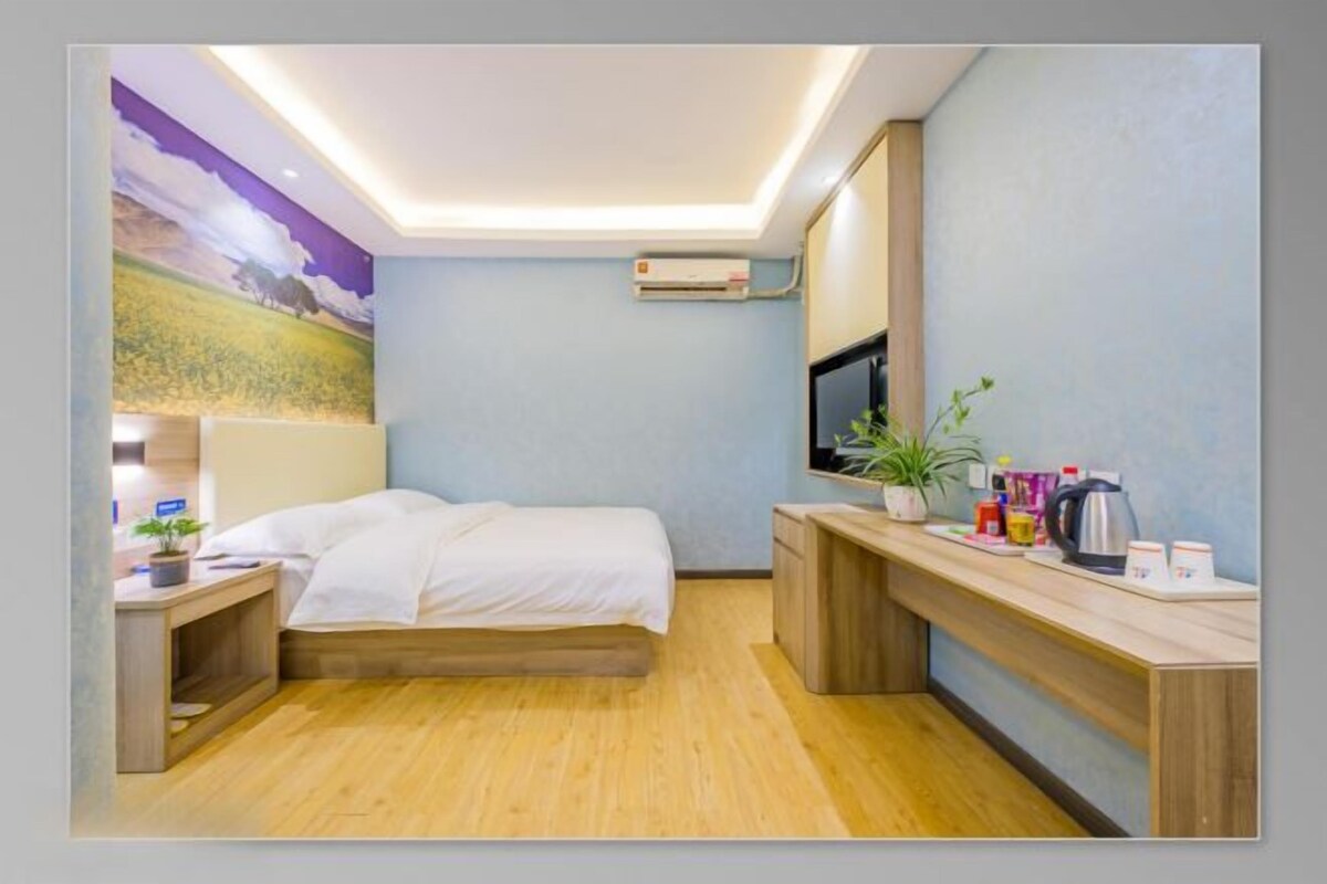 A hotel room with a bed, bedside table, a desk with a chair, a TV, and a wall-mounted air conditioner.