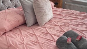 Ein ordentlich bezogenes Bett mit rosa Bettwäsche und grauen Kissen, ein kleiner Nachttisch aus Holz und ein Heizkörper in der Nähe eines Fensters mit Jalousien.