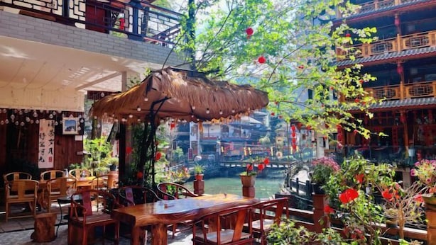 An indoor dining area with wooden furniture, a thatched roof, and a view of traditional architecture.