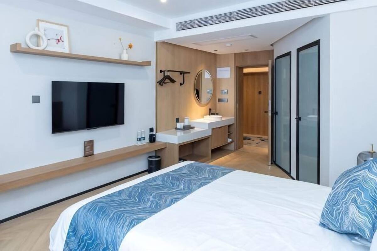 A modern hotel room with a bed, TV, bedside table, mirror, and a bathroom with a sink and shower.