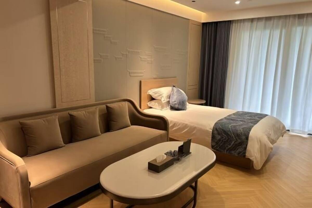 A hotel room with a sofa, a bed, a small table, and a large window with curtains.
