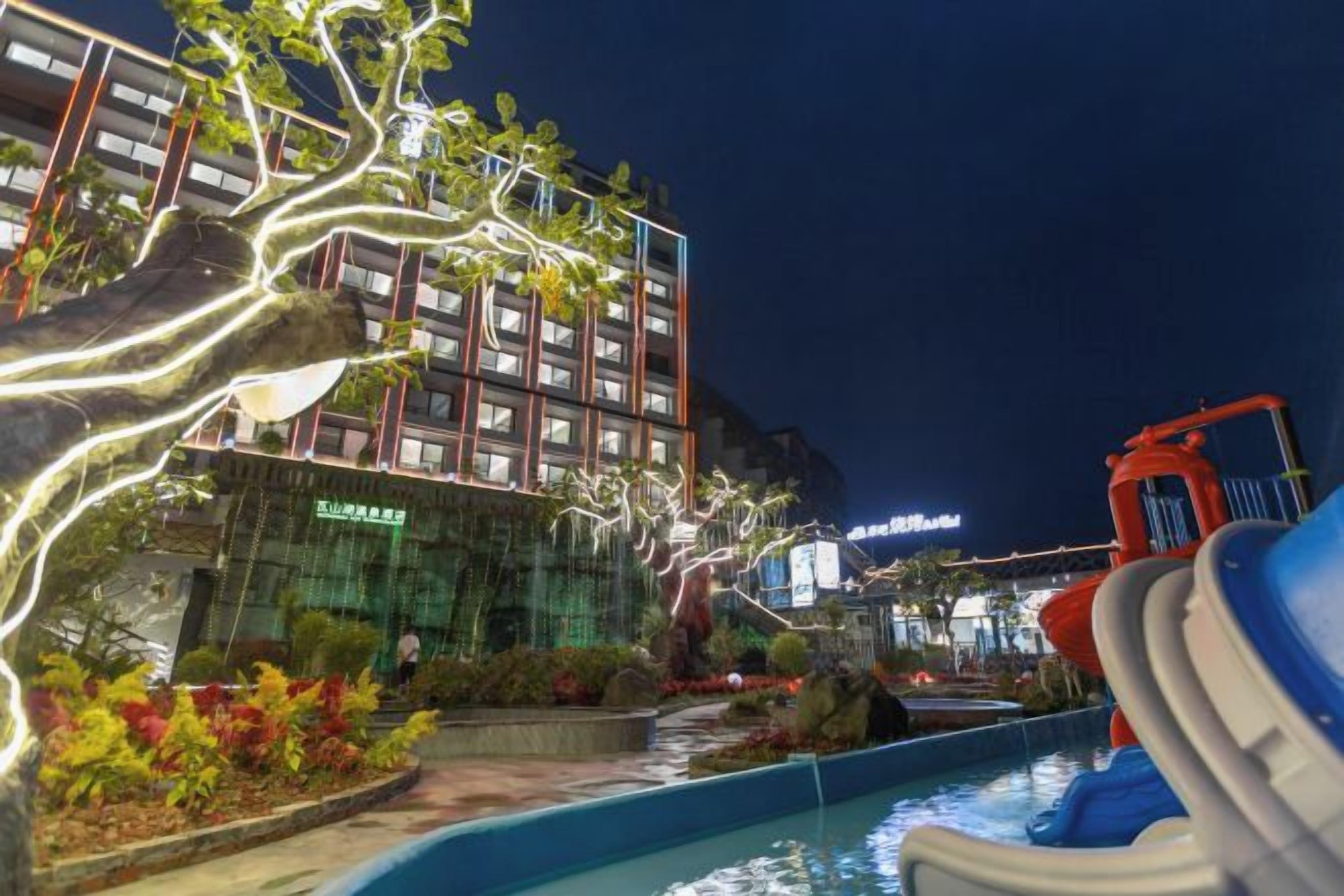 Vista nocturna de un hotel con piscina, toboganes y árboles iluminados.
