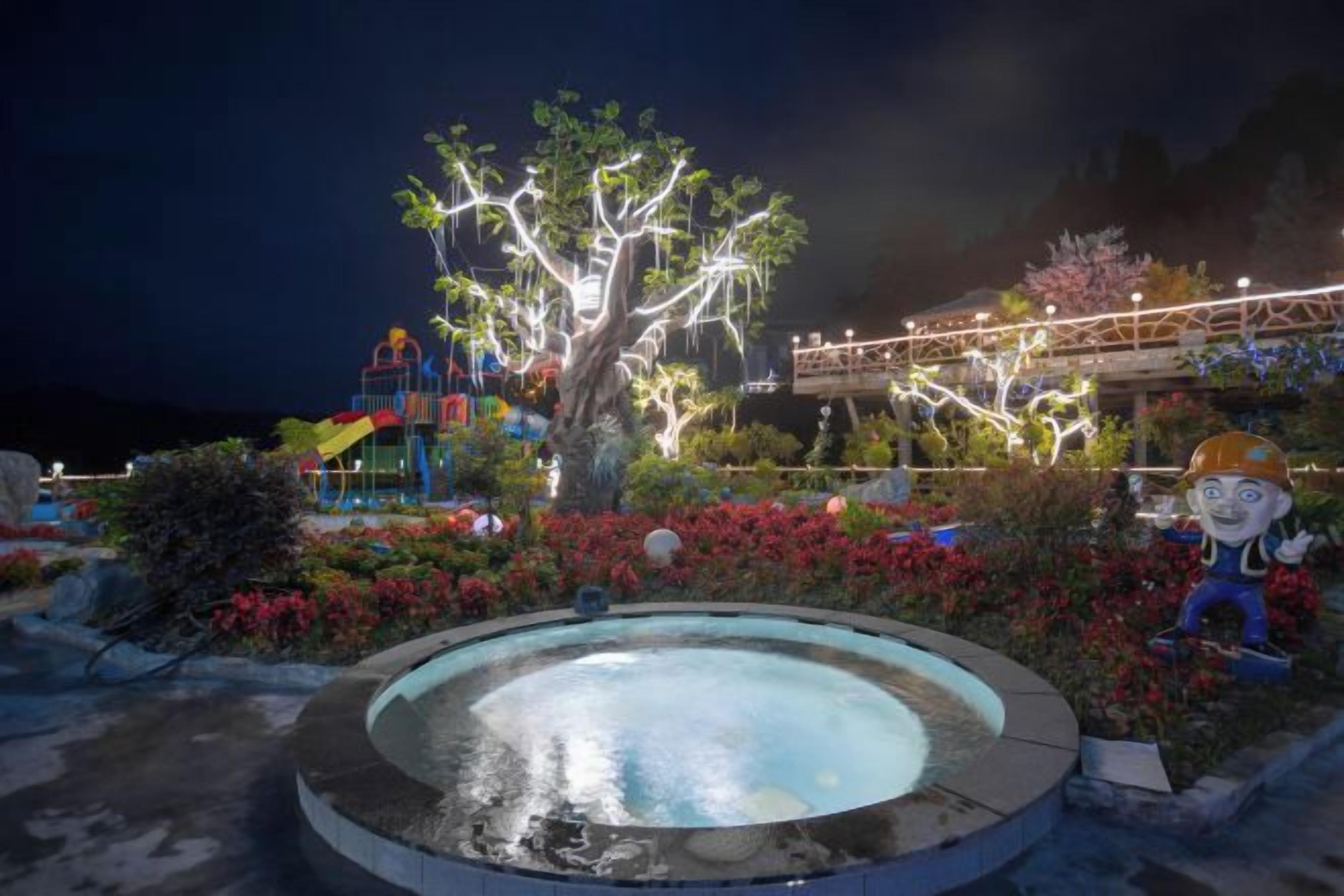 Espacio al aire libre nocturno con piscina circular, árbol iluminado y varias estructuras de colores.