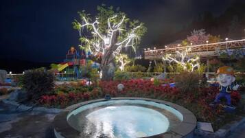 Espacio al aire libre nocturno con piscina circular, árbol iluminado y varias estructuras de colores.