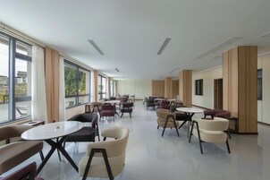 Un espacio interior amplio con diversas disposiciones de asientos, incluyendo mesas redondas y sillas, y grandes ventanales que aportan luz natural.