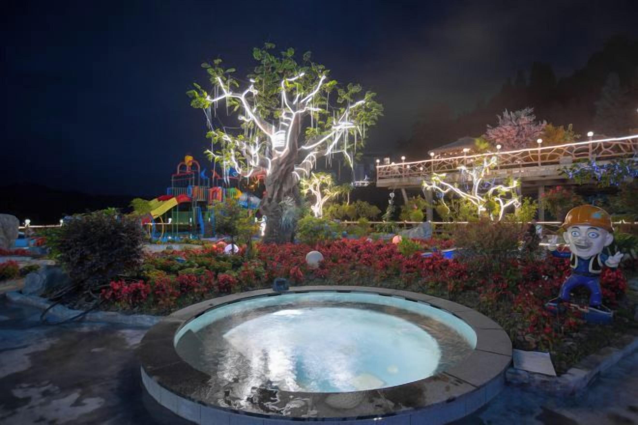 A nighttime scene with a large illuminated tree, a pool, and a colorful play structure.