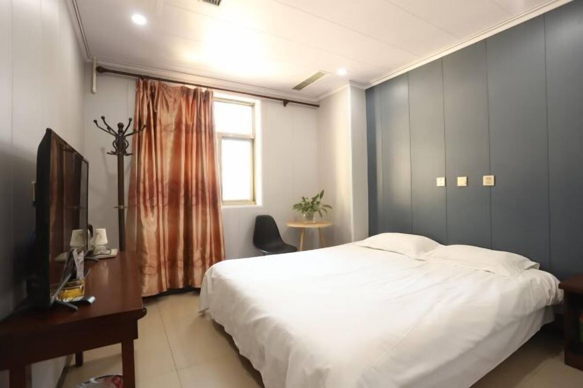 Een hotelkamer met een bed, een bureau met stoel, een kapstok, een raam met gordijnen en een kleine tafel met een plant.