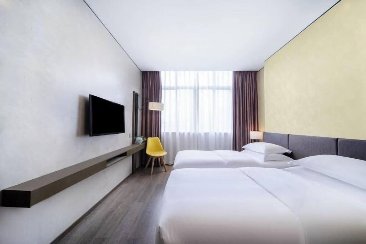 Une chambre d’hôtel équipée d’un téléviseur fixé au mur, d’un bureau avec une chaise, de deux lits et d’une grande fenêtre avec des rideaux.