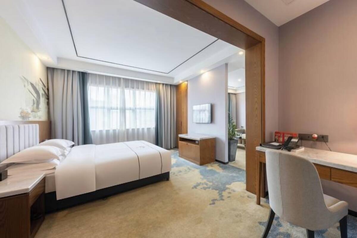 Habitació d'hotel amb llit, escriptori, cadira i una finestra gran amb cortines.
