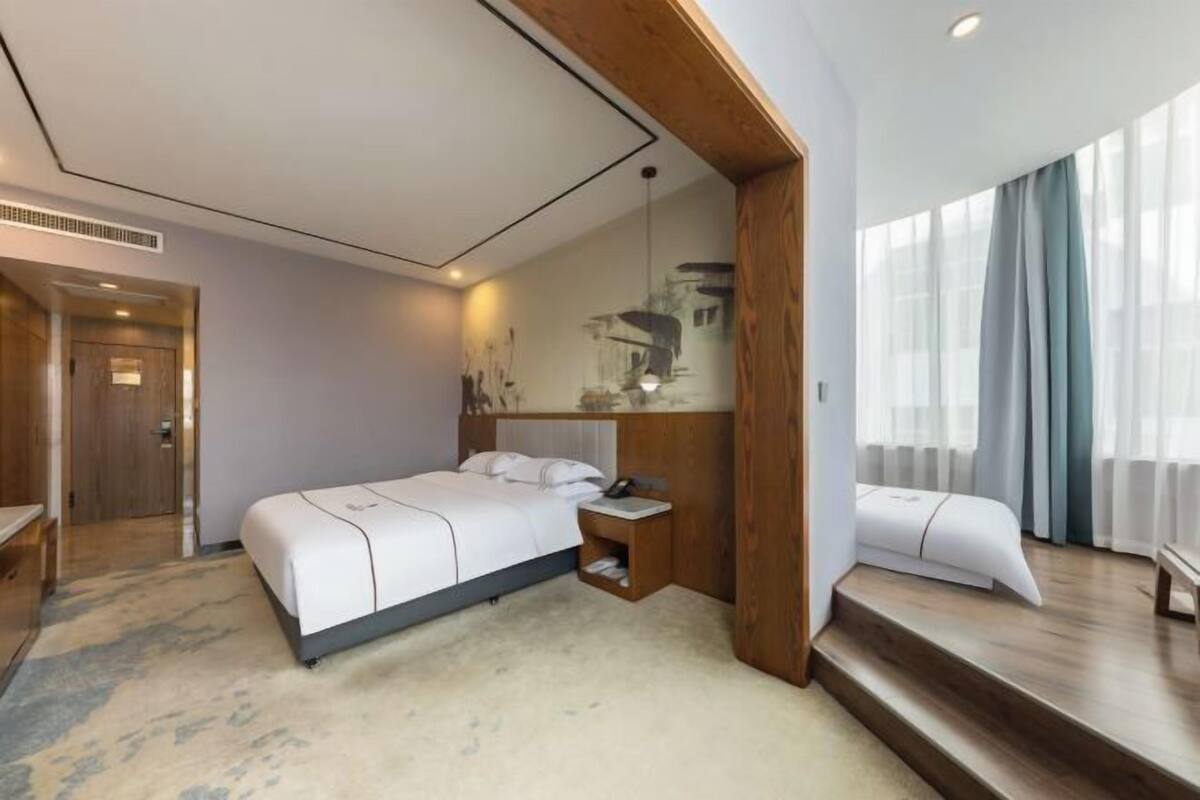 Habitació d'hotel moderna amb un llit gran, una petita taula de nit, un panell frontal de fusta i vista a la ciutat a través de grans finestres.
