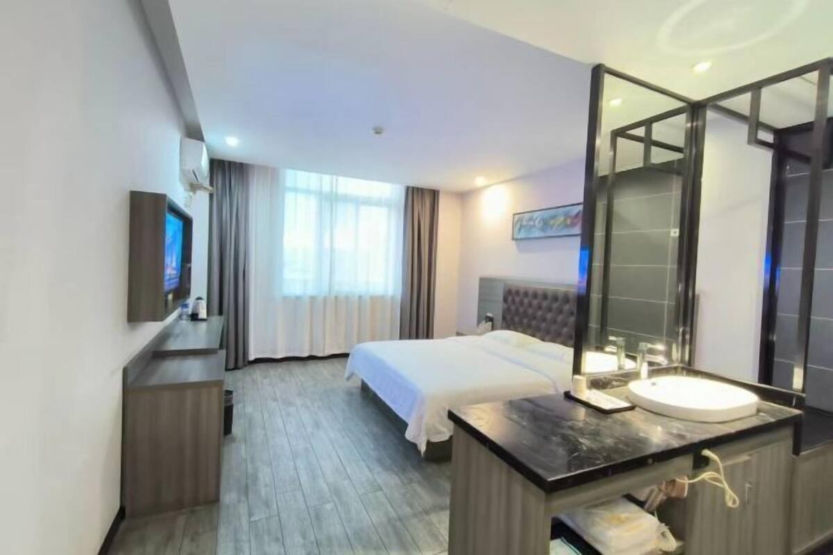 A hotel room with a bed, a bedside table with a mirror, a TV, and a window with curtains.