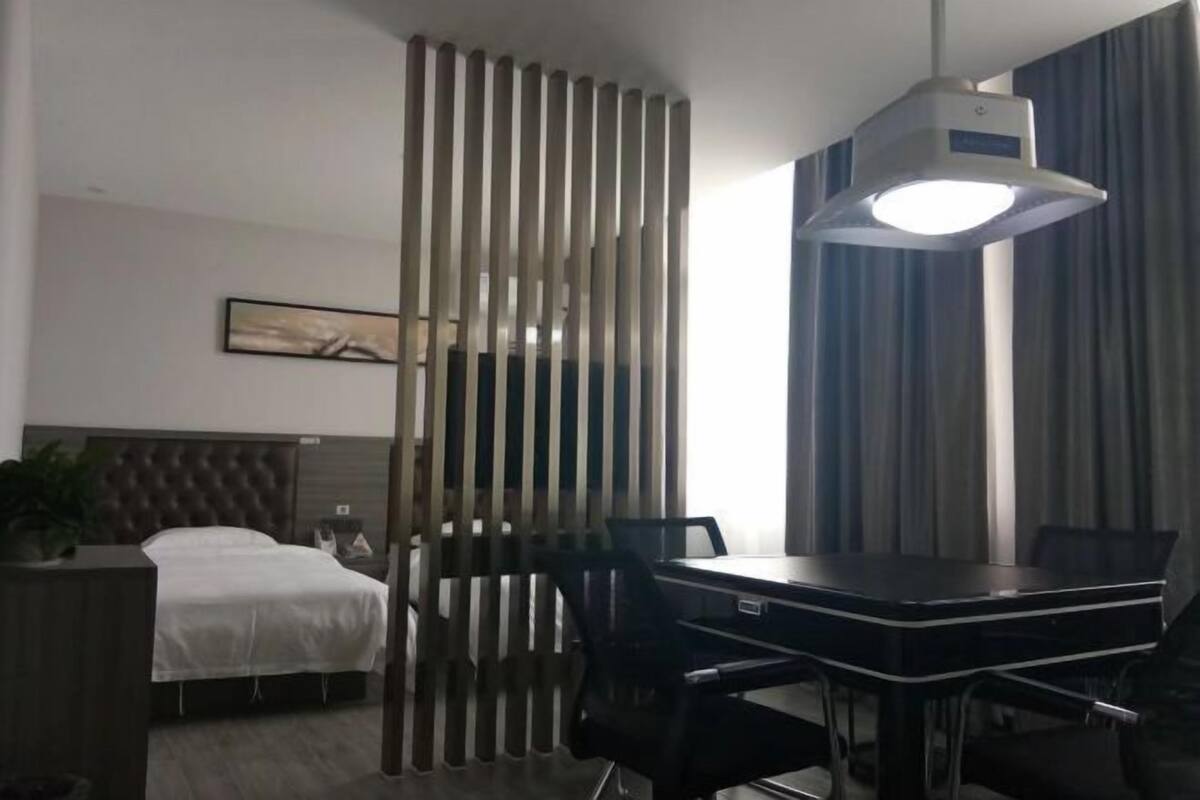 A hotel room with a bed, a desk with chairs, a large window with curtains, and a ceiling light.