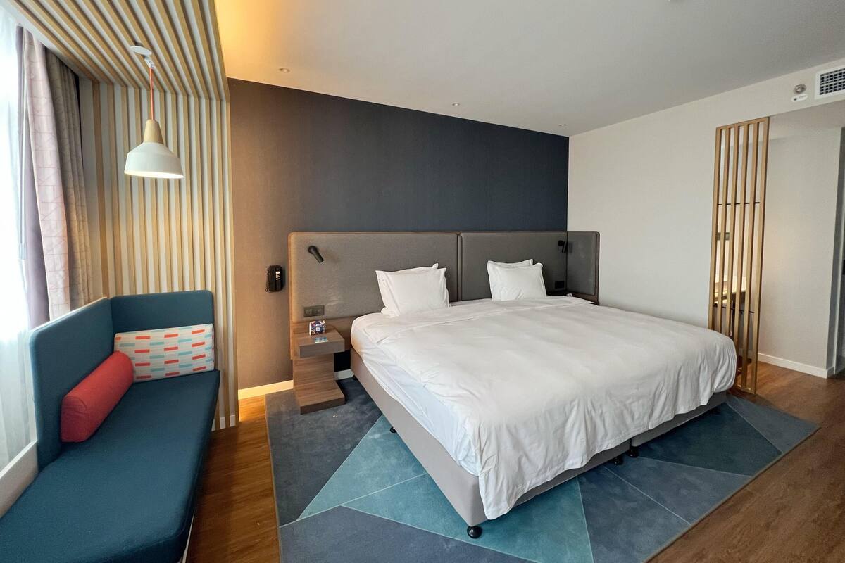 A modern hotel room with a large bed, a sofa, a nightstand, and a lamp.