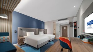 A hotel room with two beds, a desk, a chair, a TV, and a bookshelf.