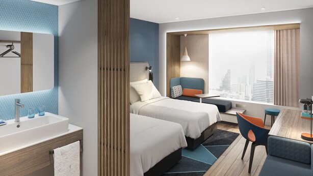 A modern hotel room with two beds, a desk, a chair, a large window with a city view, and a bathroom with a sink and mirror.