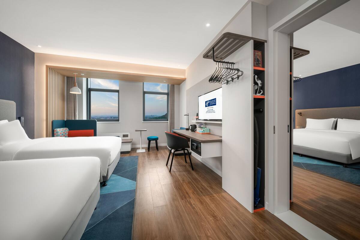 A hotel room with two beds, a desk, a chair, a TV, and a view of the city.
