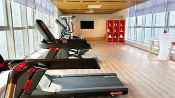 Un gimnasio con cintas de correr, un televisor montado en la pared y un estante rojo con objetos decorativos.