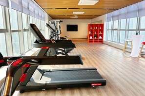 Un gimnasio con cintas de correr, un televisor montado en la pared y un estante rojo con objetos decorativos.