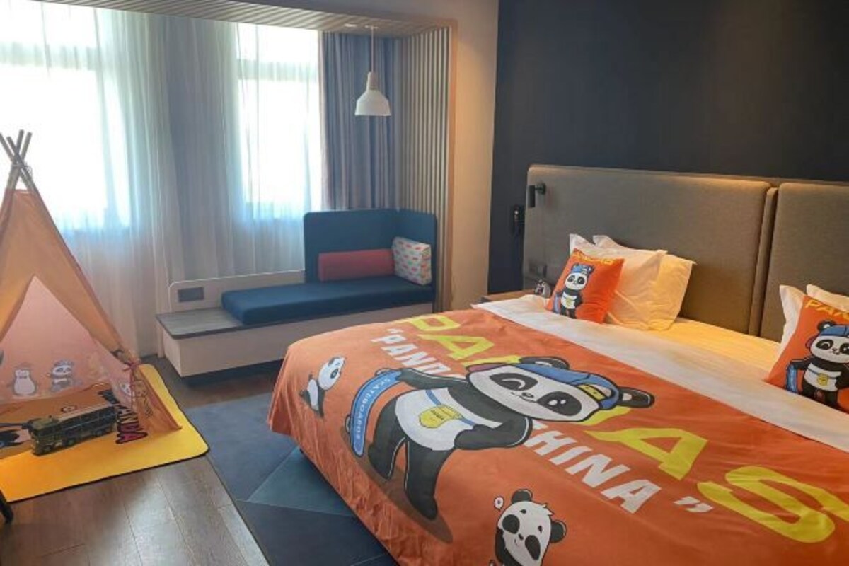 Una camera d'albergo con un letto con copriletto a tema panda, un divano con un cuscino, un tavolino con un giocattolo e una finestra con tende.