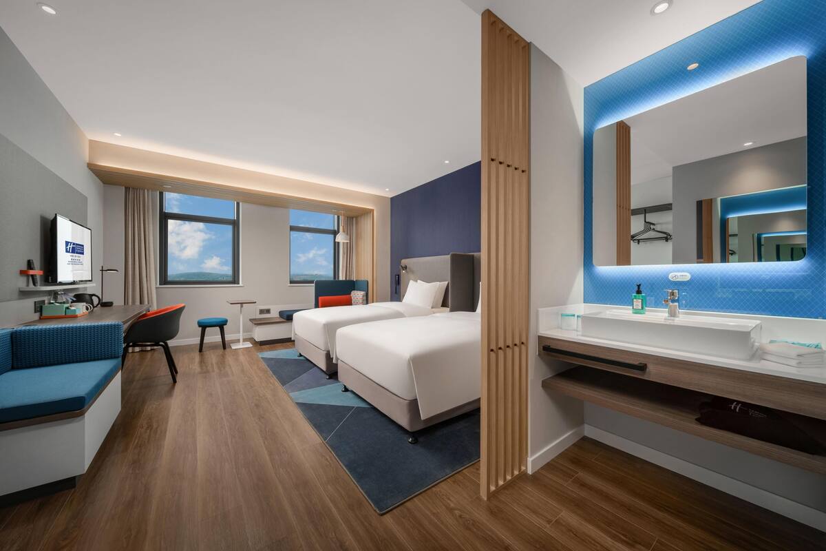 A modern hotel room with a large bed, a desk with a chair, a sofa, a bathroom with a large mirror, and a view of the sky.