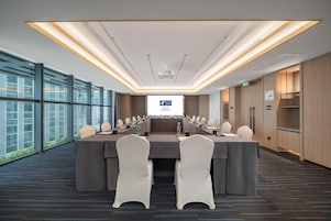 Un salón de conferencias con una larga mesa, sillas, una pantalla de proyector y ventanas amplias.