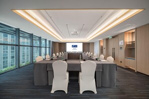 Un salón de conferencias con una larga mesa, sillas, una pantalla de proyector y ventanas amplias.